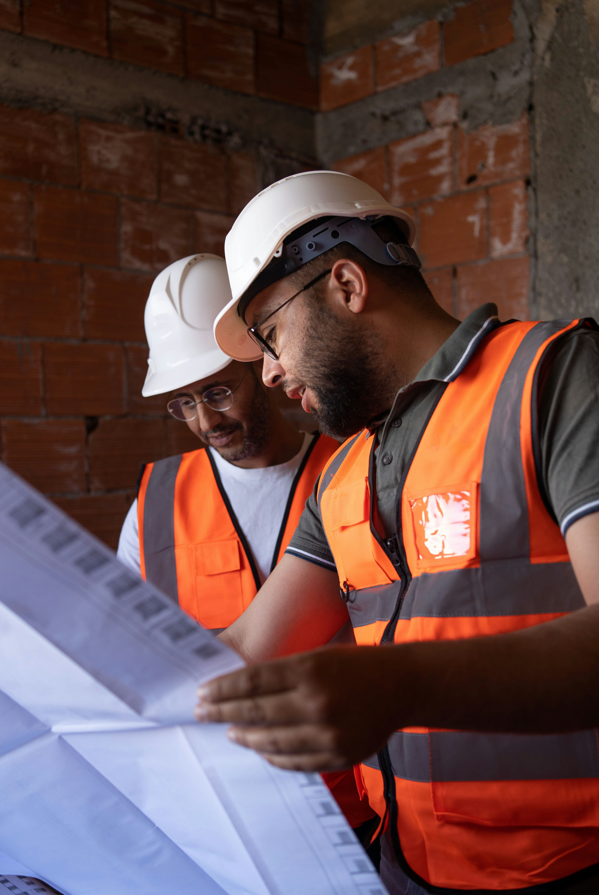 Deux travailleurs en chantier portant des casques de sécurité blancs et des gilets orange, examinant des plans de construction.