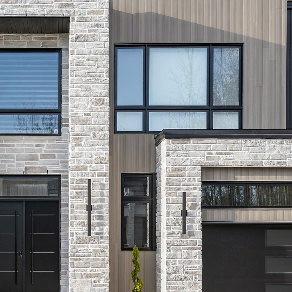Façade moderne d'une maison avec des murs en pierre grise, fenêtres noires et porte de garage assortie.