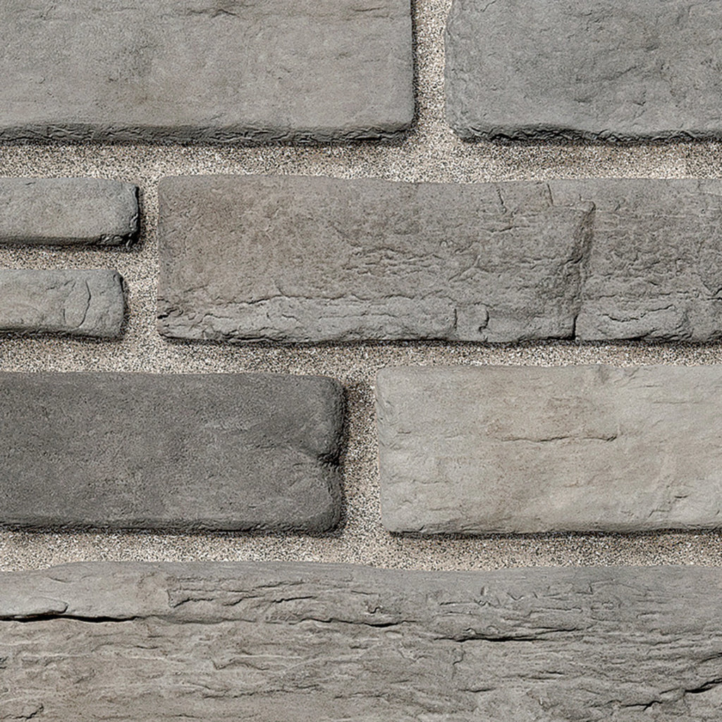 Briques en pierre grise de différentes tailles, disposées en motif sur un mur, avec un fond de sable.