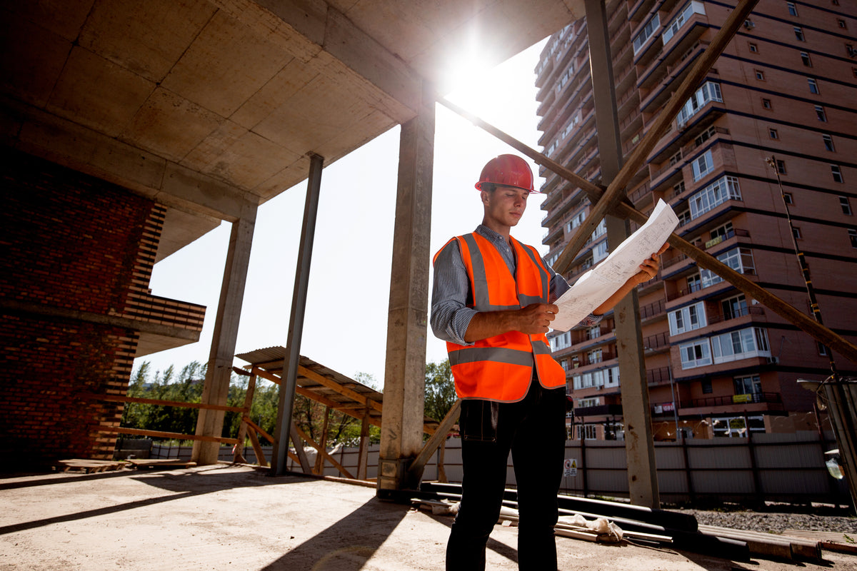 Homme en gilet de sécurité orange et casque rouge, tenant des plans, sur un chantier de construction avec des bâtiments en arrière-plan.