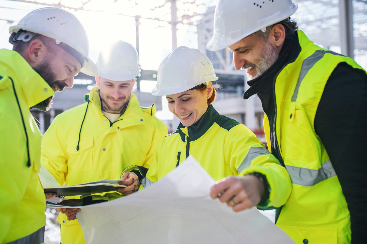 Quatre travailleurs en casques de sécurité et vestes jaunes fluo examinant des plans sur un chantier de construction.