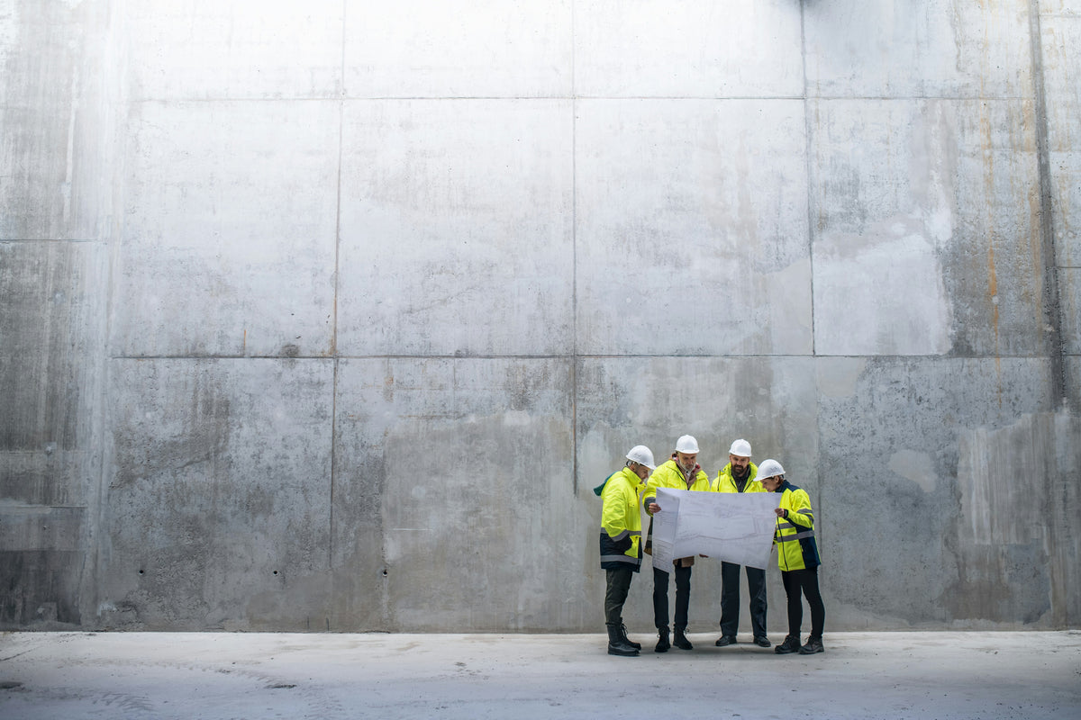 Quatre travailleurs en chantiers portant des vestes jaunes et des casques blancs, examinant des plans sur un mur de béton.