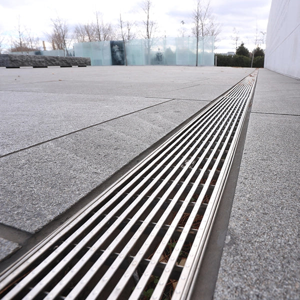 Grille de drainage en acier inoxydable sur un sol en dalles grises, avec des arbres en arrière-plan et un ciel nuageux.