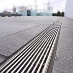 Grille de drainage en acier inoxydable sur un sol en dalles grises, avec des arbres en arrière-plan et un ciel nuageux.