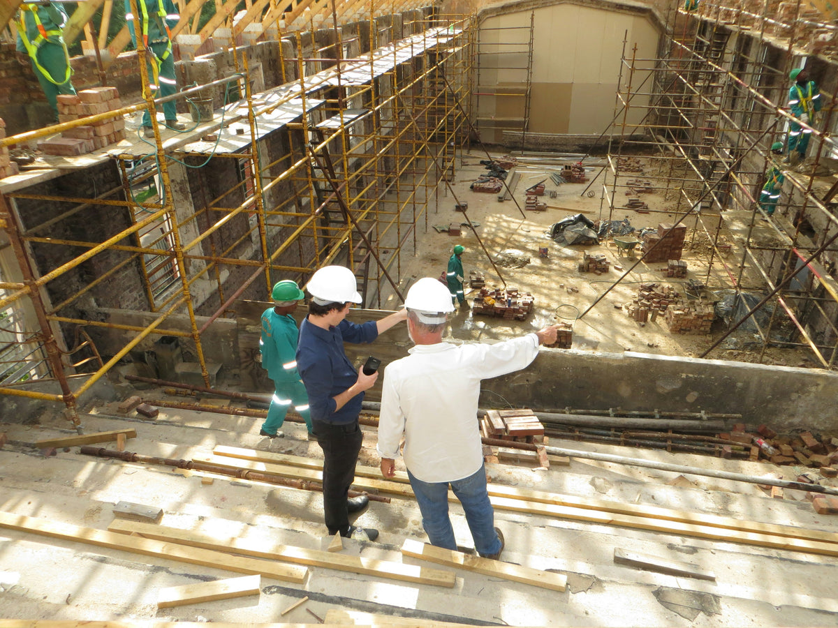 Deux travailleurs en casques de sécurité discutent sur un chantier de construction avec des échafaudages et des matériaux de construction visibles.