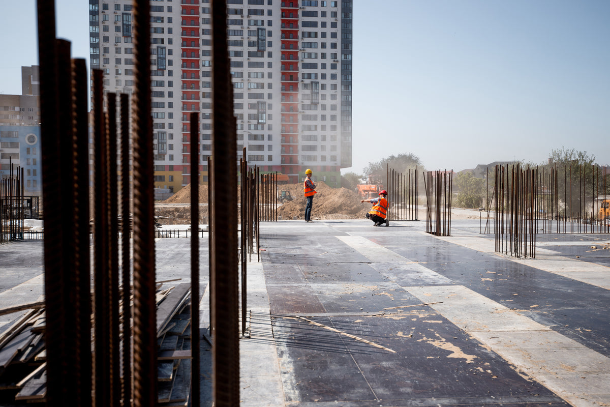Chantier de construction avec deux travailleurs en gilets de sécurité, armatures en acier et bâtiment en arrière-plan.