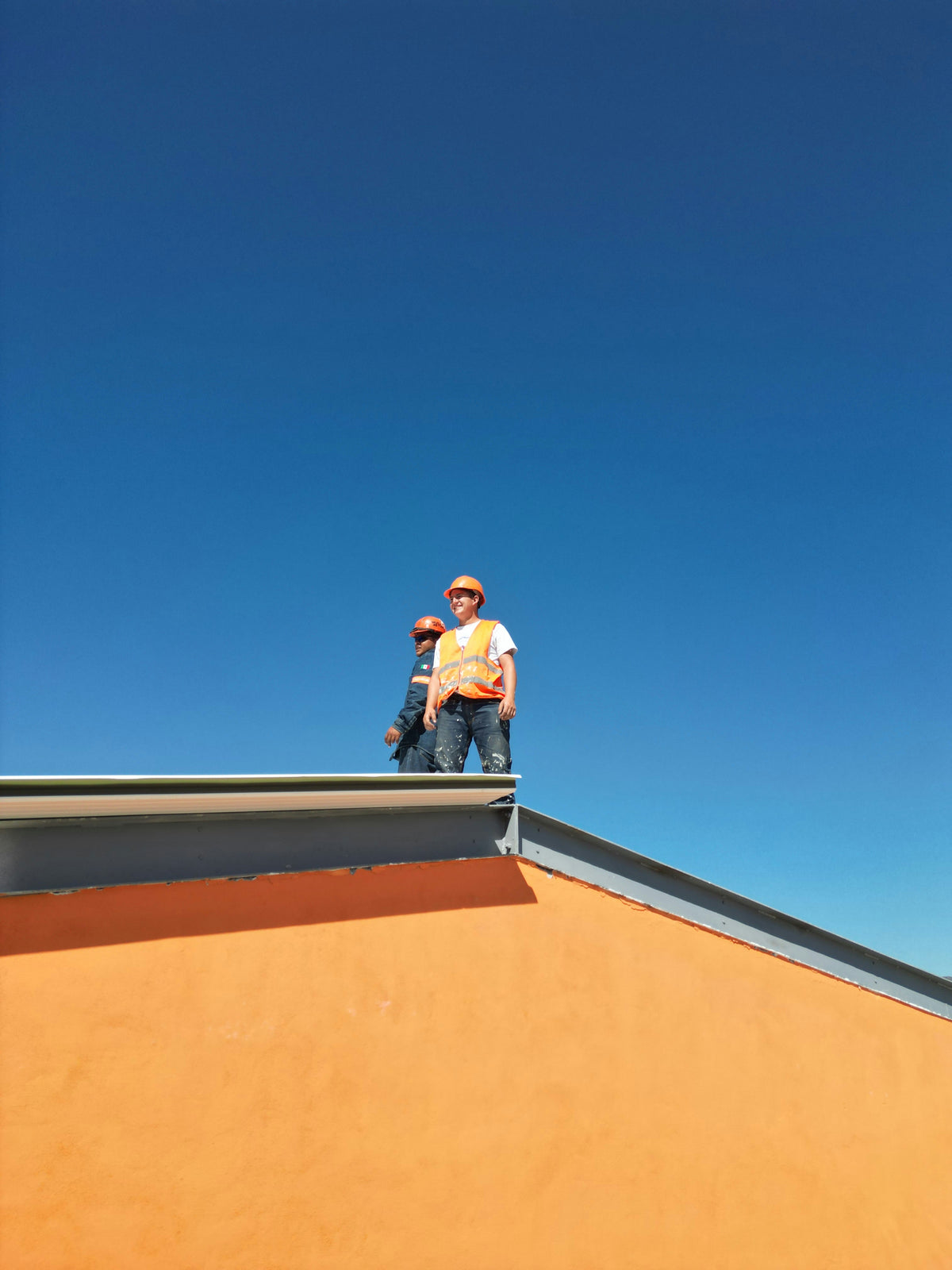 Deux travailleurs casques et gilets orange sur le toit d'un batiment.